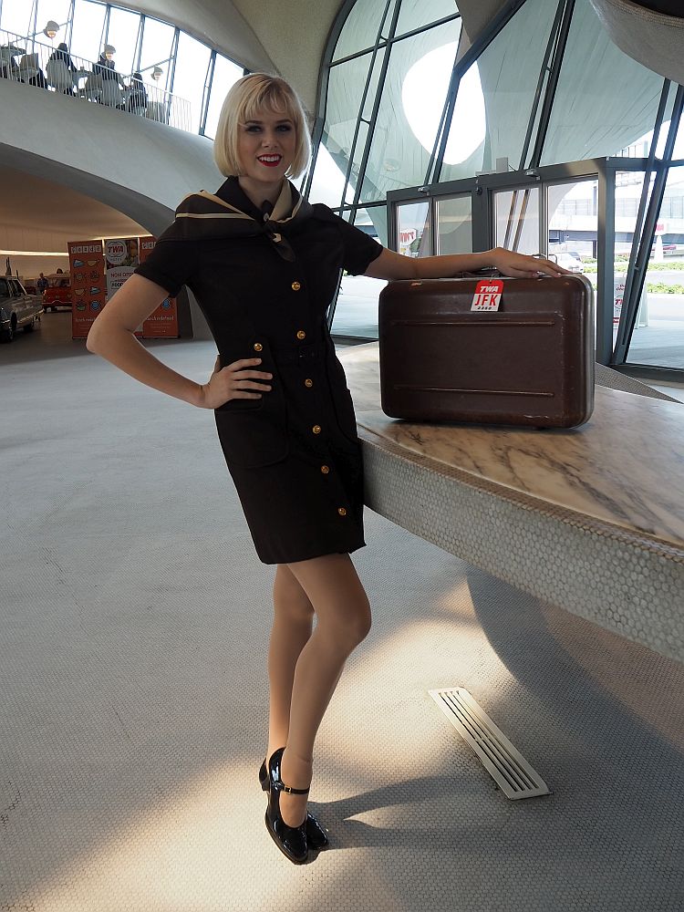 The attendant's outfit is brown and close-fitting, with a mid-thigh-length skirt and short sleeves. She wears black pumps and leans on a small suitcase.