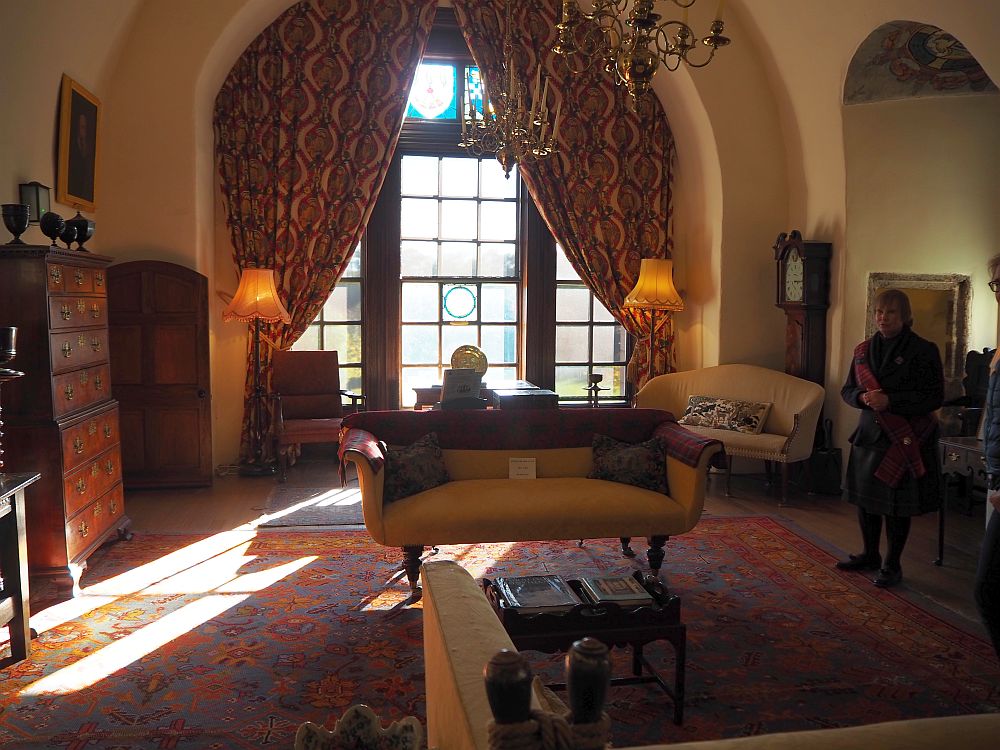 An arched window with draped curtains lets light into a room with a couch, a dark carpet, armchairs, cupboards, etc.