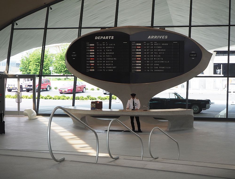 Inside the front entrance of the TWA Hotel, an actor dressed as a pilot poses underneath the departures board. A vintage car is parked outside. 