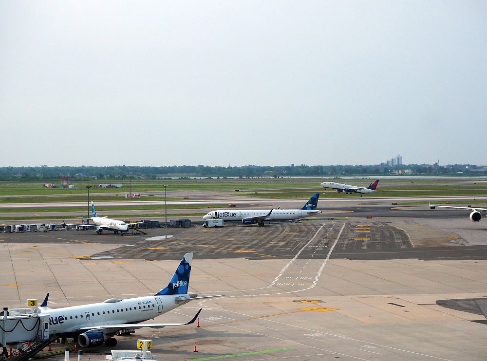 In the foreground left is a JetBlue plane parked at a Terminal 5 gate. In the middle distance, a plane stands waiting, and a bit further away, a plane is just lifting off the ground.