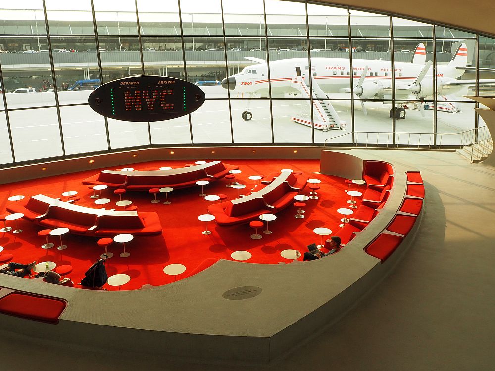 The lounge is carpeted in bright red, dotted with small white tables and red-upholstered benches. Light enters through a wall of large windows, behind which the Connie is visible.