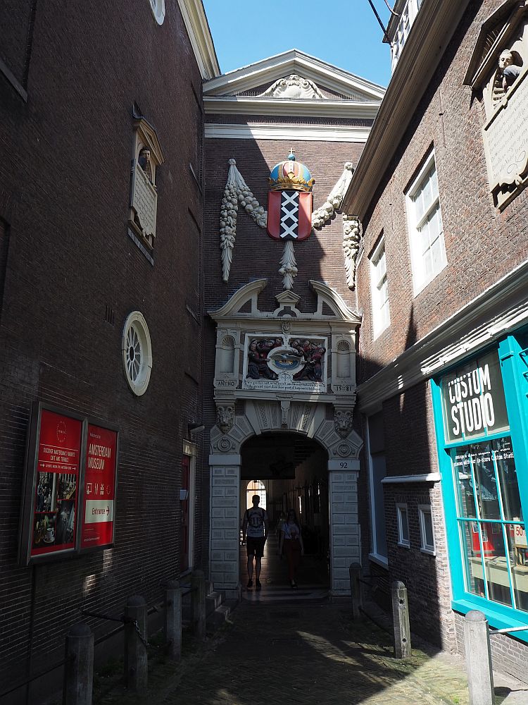 Down a narrow alleyway, a gateway is visible that is the entrance to the Amsterdam Historical Museum. It is arched and rather ornate, with a distinct lean to the right.