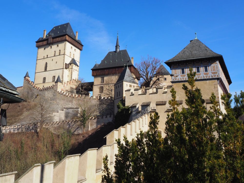 This view from one end of the castle shows three towers, the tallest of which is the Great Tower. The walls are off-white and the roofs are grey. Visiting Karlstejn Castle from Prague