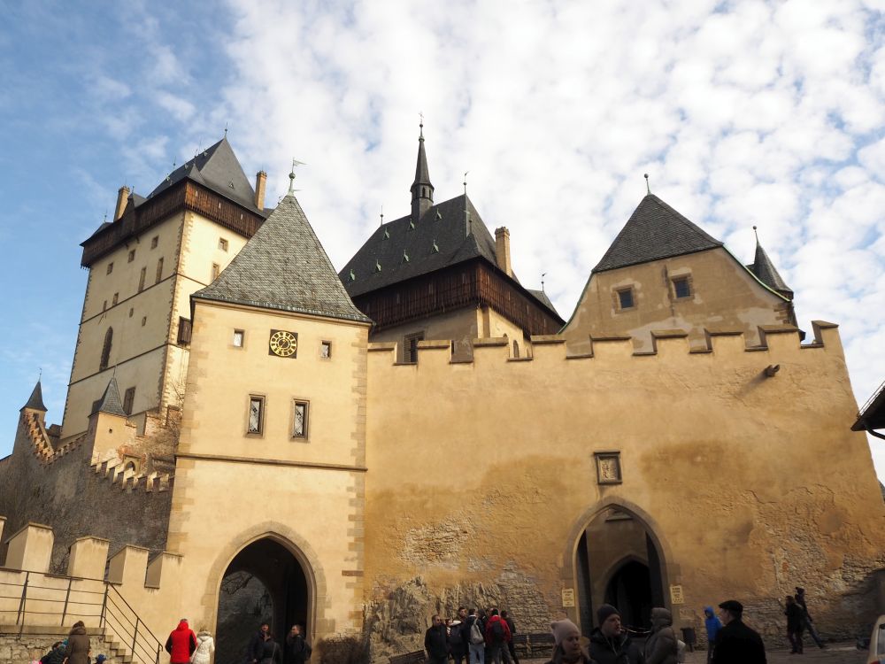 A large wall with crenellations and a gateway in its middle, and three towertops visible behind it. 