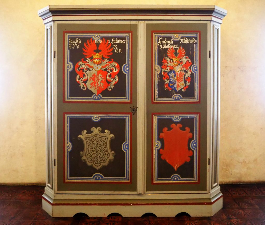 The cabinet is painted in bright colors, with four shields depicted: coats of arms, perhaps. The two top ones seem complete while the two lower ones are only outlines.