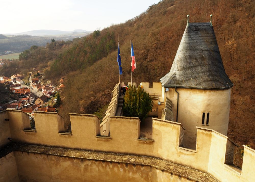 a piece of the crenellated wall and a small tower  with a steep gray roof in the foreground, a valley between hills in the background, with a cluster of houses.