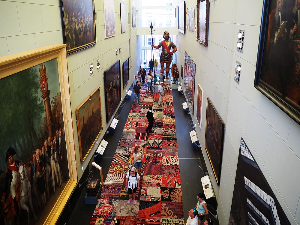 Looking from the second floor down the length of the central hall of the Amsterdam Historical Museum. The hall is lined with huge paintings and a very large statue of a warrior of some sort holding a spear stands at the far end.