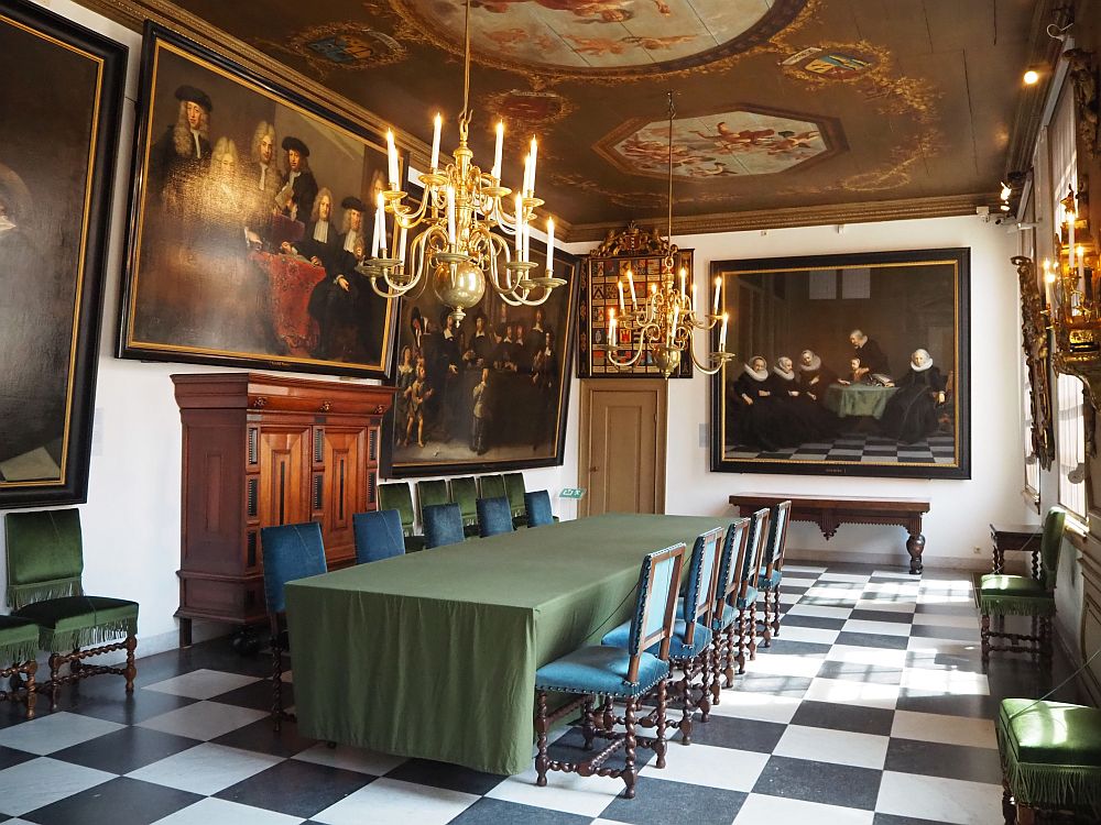 The regents' room at the Amsterdam historical museum is quite grand, with a table down the middle and a chandelier above it. The walls hold large paintings of regents.