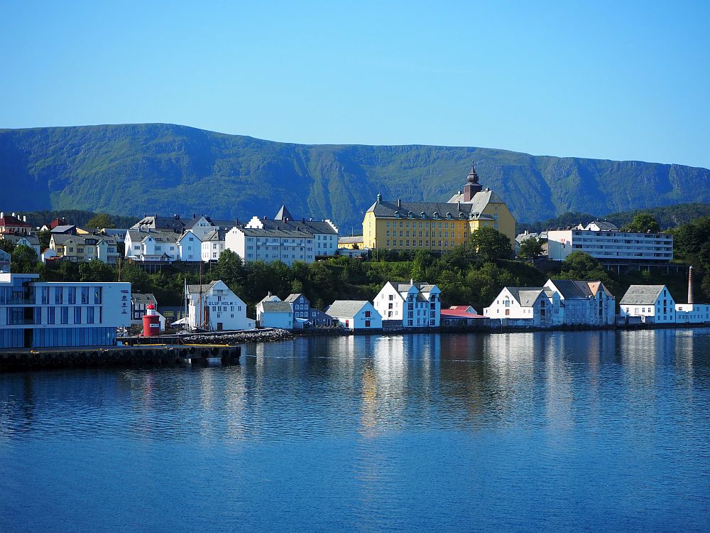 Two lines of buildings as seen from the water. One row sits right at the waters edge, while the other sits on a flat-topped rise above them. One large building, painted yellow, looks like some sort of school or museum in a 19th century style.