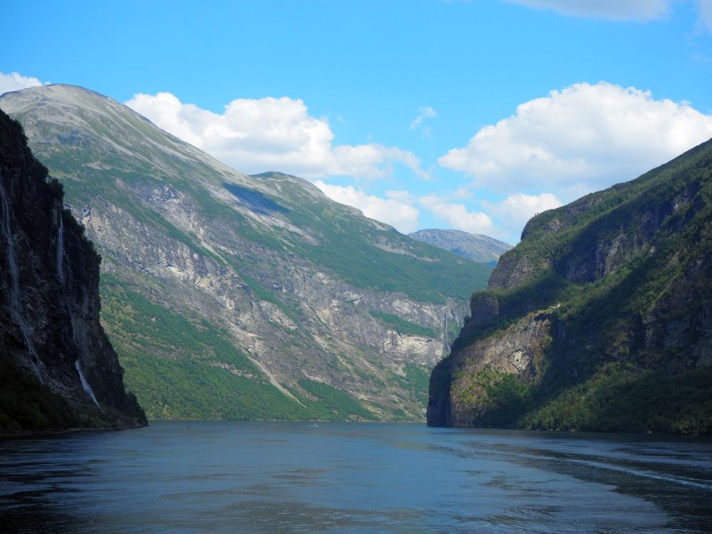 A view into the fjord: dark water ahead, a steep cliff on either side and a larger mountain straight ahead with a mix of rock and green ground cover.