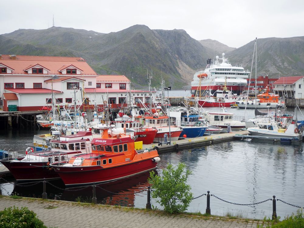 A cluster of small fishing boats moored on a dock, with the Hurtigruten ship visible in the background, and rocky, bare mountains visible beyond that.
Hurtigruten review.