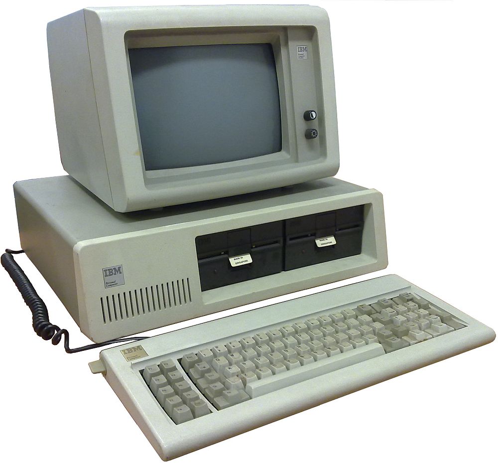 An early IBM personal computer, beige, with a fat monitor sitting on the computer itself, which has two slots for floppy disks. A keyboard sits in front, not much different from keyboards today.