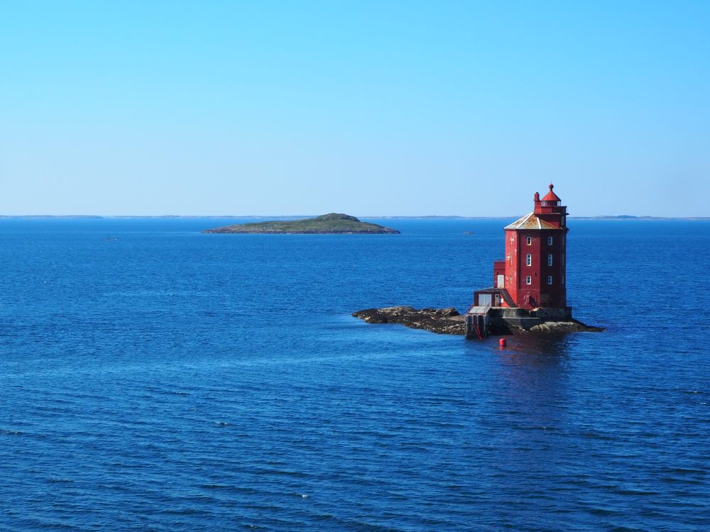 A red lighthouse on a very small, rocky bit of land, surrounded by blue sea, with another rocky island in the background.