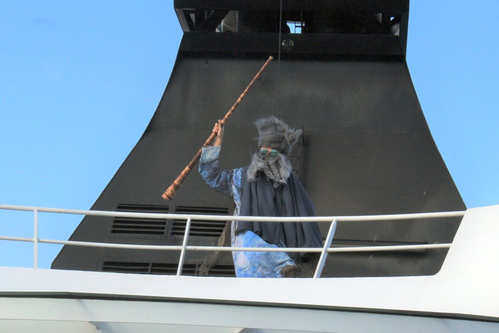 A person standing on the top deck above the photographer, dressed in a blue robe with a black cape, wearing a large fake gray beard and hat and sunglasses, waves a staff.