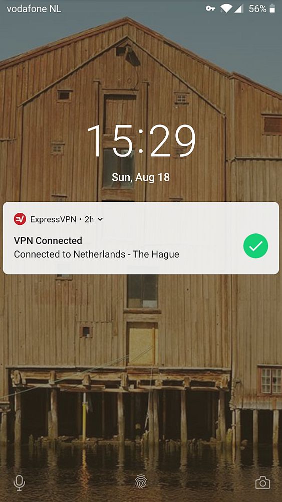 A screenshot of a telephone start screen. Behind, a photo of an old, brown, warehouse. In the middle: the time, the date, and a message saying "Express VPN. VPN Connected. Connected to Netherlands-The Hague"