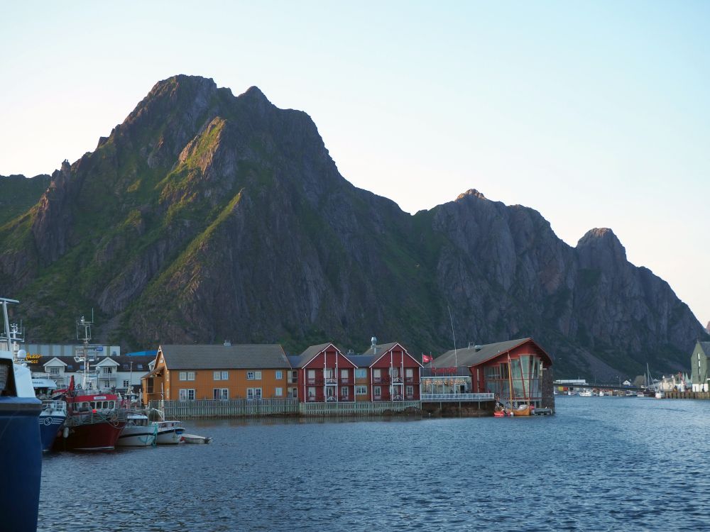 A line of buildings on a pier with peaked roofs in orange and red. Very rocky mountain behind.