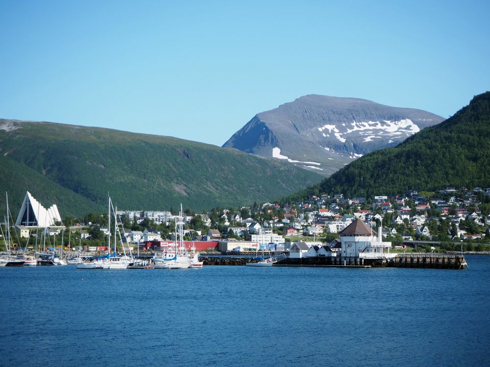 A cluster of buildings, with a large, modern church on the left and sailboats moored in front. Behind, two green hills and then, behind them, a higher hill with snow in patches on its side.