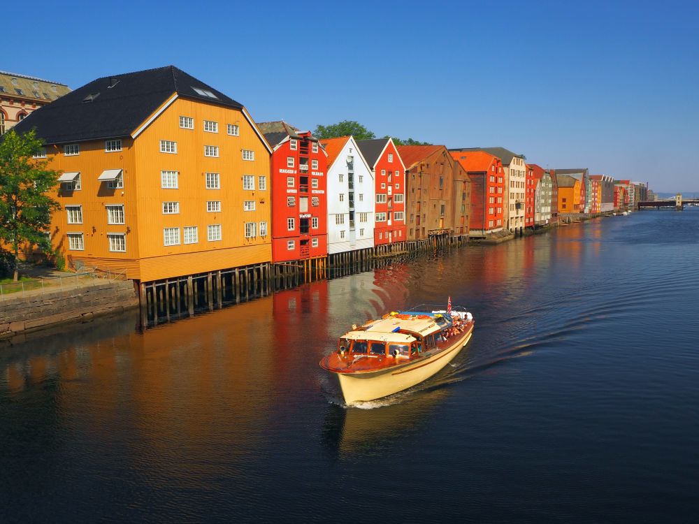 A row of large former warehouses extend over the shore of a river on stilts. Each is painted in yellow, red, white or brown. A small tour boat passes in front, in the foreground of the photo.