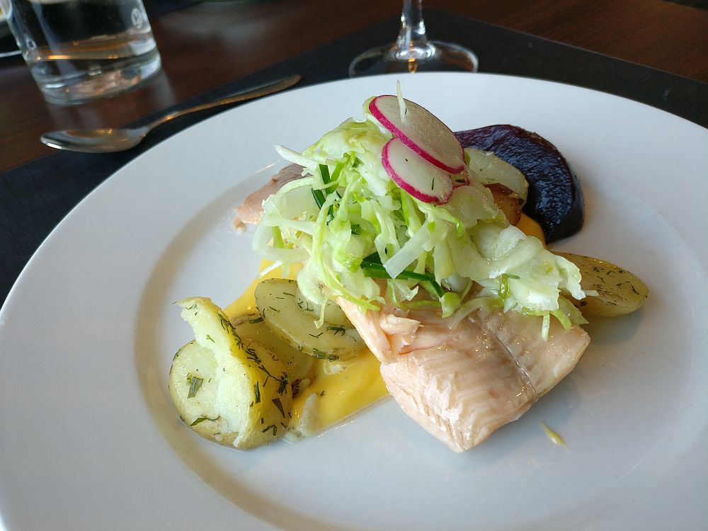 The food is presented in a pile on a white place: first the hollandaise sauce underneath, then dill potatoes are visible on either side, under a piece of light-pink fish, which in turn has a small amount of shredded cabbage and sliced radish on top. The beet is only partly visible behind the fish.