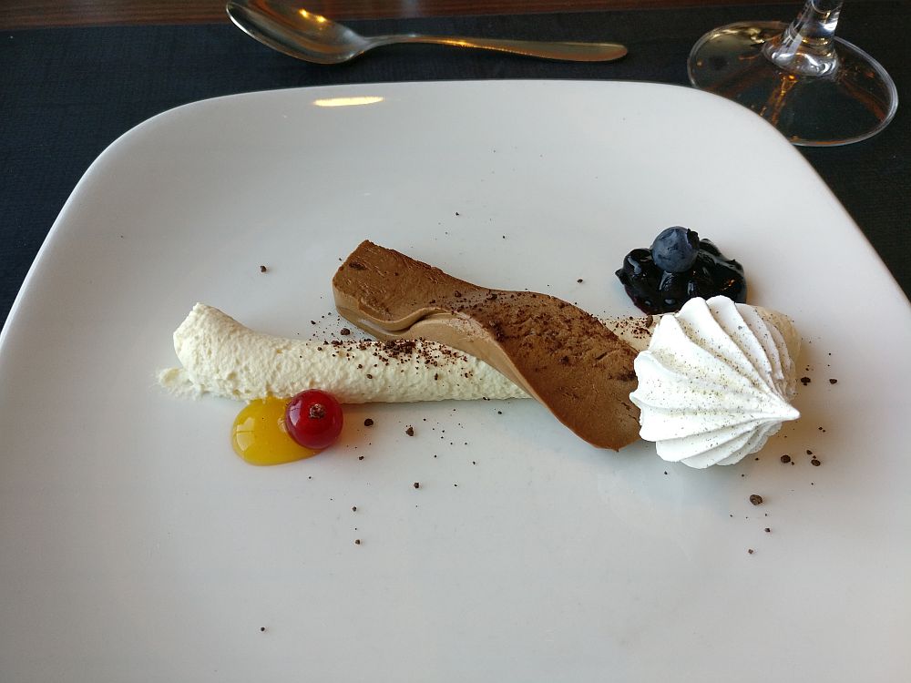 A white cream lies horizontally in a line across the plate. A piece of chocolate mousse lies across that. On the side are a bit of blueberry compote, a white meringue, and a red berry (lingonberry?) on a dab of yellow sauce.