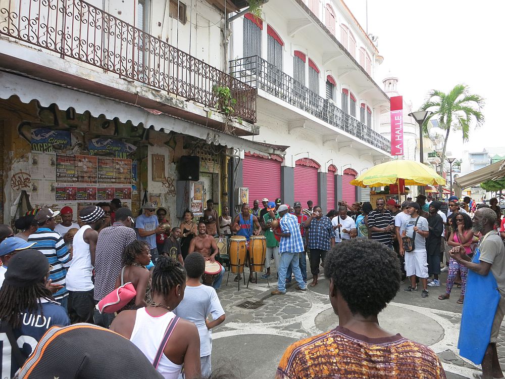 On a city street, closed storefronts on the left of the picture, with lacy iron railings above, the drummer sit under the balcony. One man is shirtless and sitting, while several others are standing and playing various drums. A crowd stands/dances in a circle around them. Judging by their dark skin, these are all locals.  10 reasons people get addicted to travel.