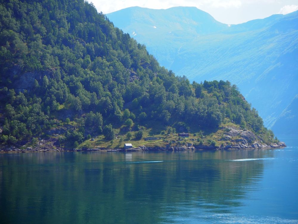 A slope of tree-covered land from top left to lower right. At its base, on the edge of the water, a tiny cottage. Huge mountains, much lighter-colored, in the background.