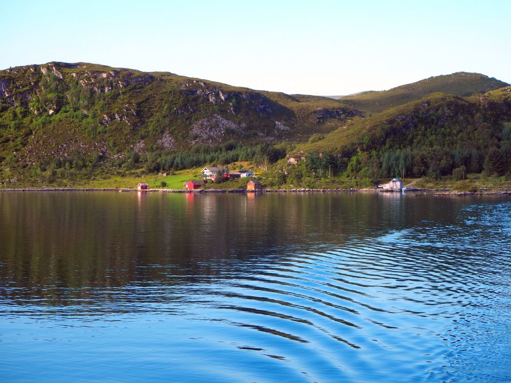 water in the foreground, gently rolling hills in the background, partly covered with trees. In a flat area between two hills is a cluster of houses: just a couple, and a couple of boathouses below them, right on the edge of water.