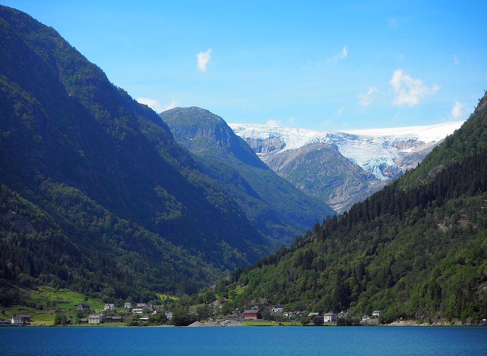 At the bottom, blue water. On the shore, a small village in a valley between very high mountains left and right. In the background are more mountains, and the furthest, tallest one is topped in snow and ice.