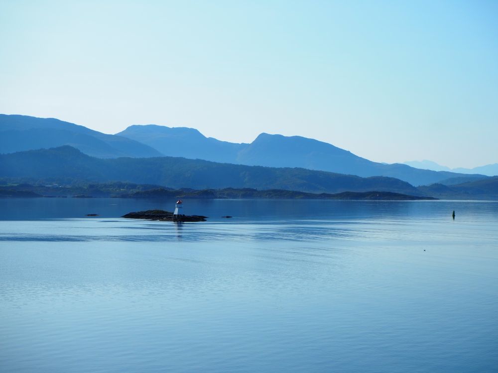 Smooth blue water. A small rock with a marker buoy on it. Behind that, rows of mountains, one behind the other. All seem blue, but darker blue in front shading to lighter blue behind.