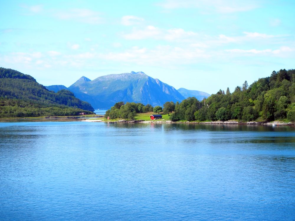 Water in the foreground, gently sloping land in the distance, covered in trees except for a cleared green field. In the field, a small red house. Behind, in the distance, a high, rocky mountain.