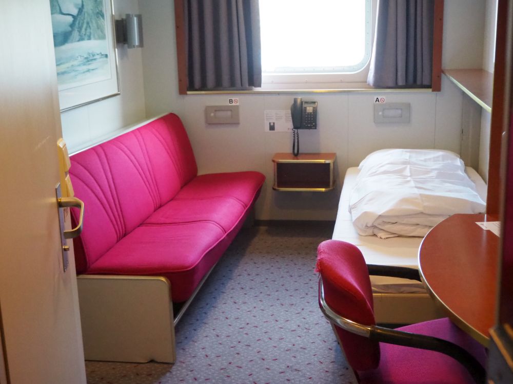 red sofa on the left, single bed on the right. A large window on the back wall and a desk and desk chair in the foreground.