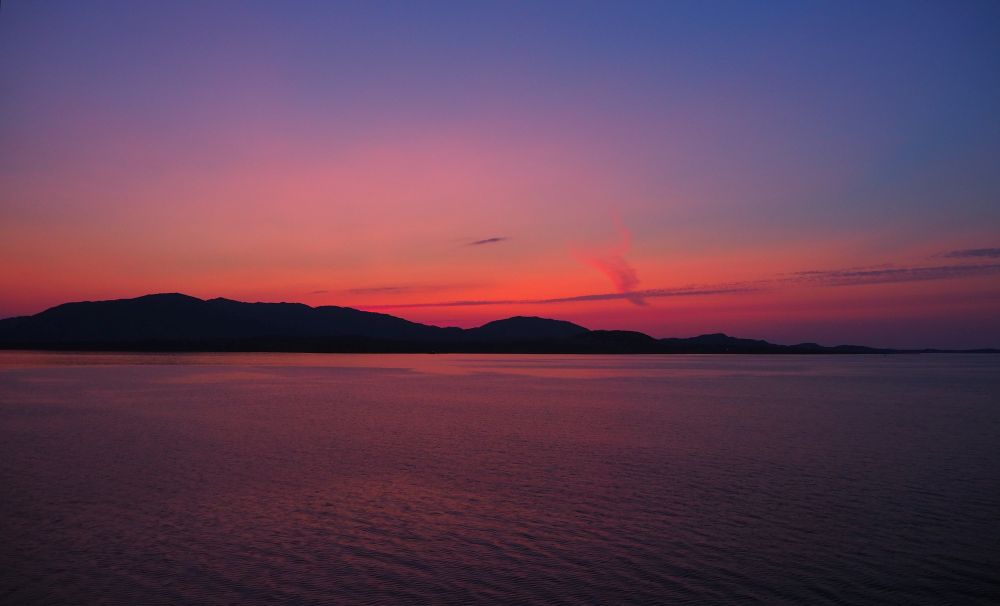 A pink and purple sunset over distant low hills, water in the foreground, very calm, reflecting the purple.