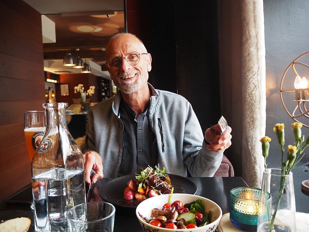 Albert, wearing a gray sweatshirt, smiles at the camera, holding his fork in one hand. In front of him is a dark brown plate with a pile of vegetables and meat in the center. In the foreground, a white bowl with salad.