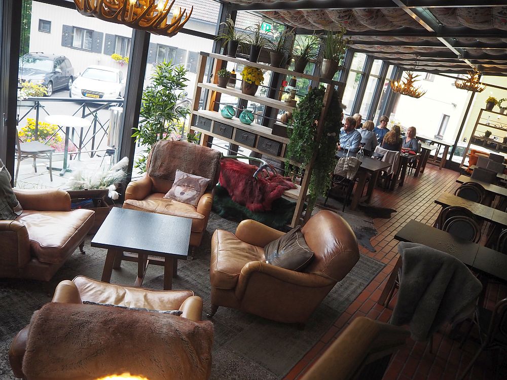 In the foreground, a cluster of soft brown armchairs with cushions. In the background, tables with people sitting at them. A brown shelving unit divides the two spaces, and big windows let in lots of light.