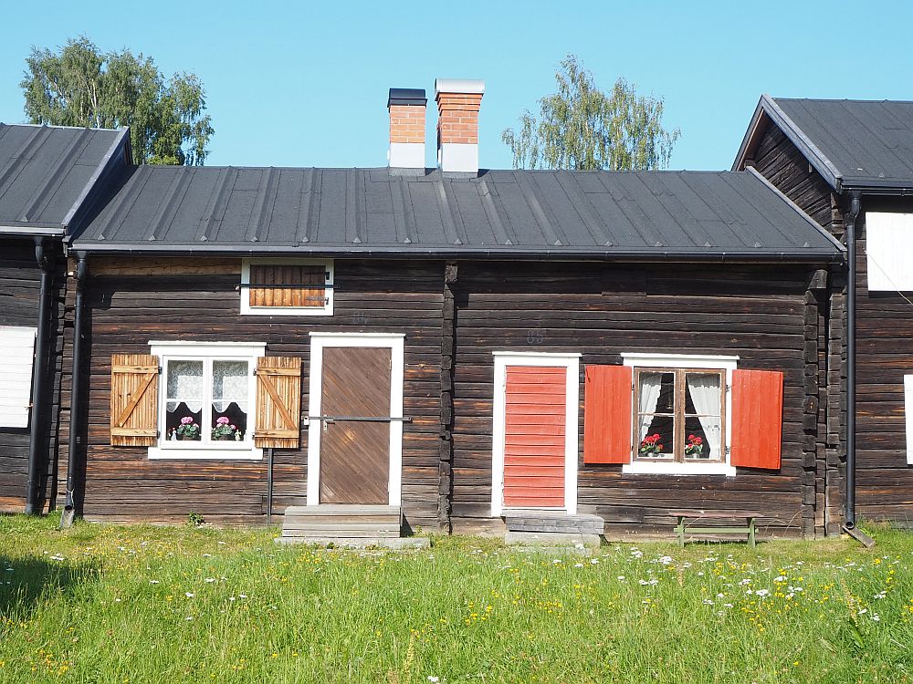 Two small houses fill the photo, with a bit of green grass in front. Each has one door with one window next to it. The left-hand one is brown with white-trimmed windows and brown shutters and door. The right-hand one is the same but the shutters and door are painted an orangy-pink color. 