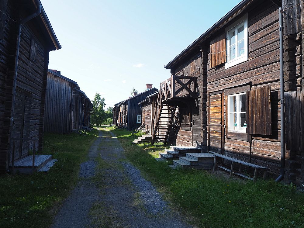 A view down a Bonnstan road. On either side: dark brown houses of flat-cut logs. Windows are trimmed in white, with brown shutters. Some houses are single-story, some are two-story. One house, on the right, has a staircase crossing the front to the upper floor.