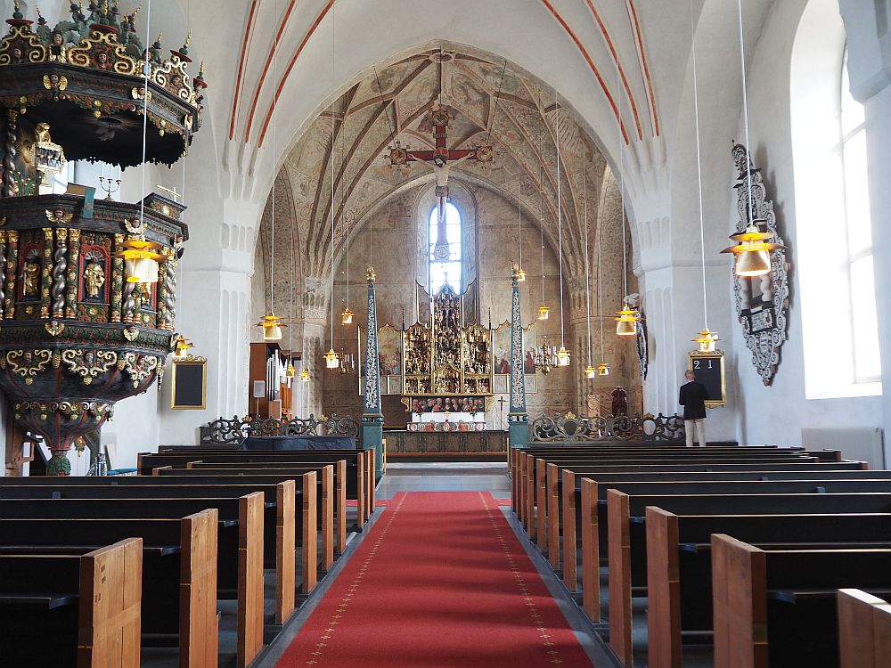 Looking down the red-carpeted aisle of the church in Gammelstad church town, rows of pews are visible on either side. At the end of the building is the altar topped with an elaborate altarpiece. A window lets light in above the altar. ON the left, an elaborate-carved pulpit is visible. 