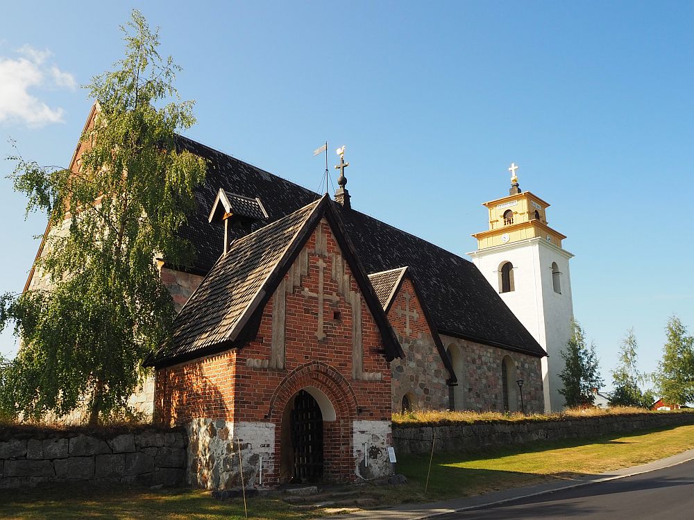 The stone church in Gammelstad church town. An archway leads to it in the foreground and a white bell tower is visible behind it.