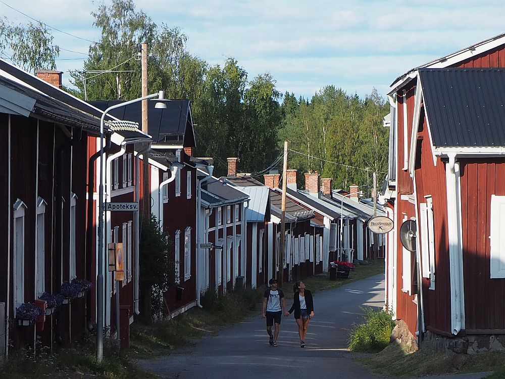 A young couple holding hands walks toward the camera along a road in Gammelstad church town, lined on both sides with small red houses, all very close together. The road turns out of sight to the right behind them.