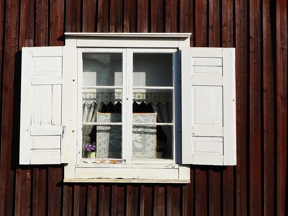 A detail photo of a window in Gammelstad church town. It has 6 panes in two columns of 3 rows. Trimmed in white, white shutters stand open on either side, bright against the dark red wall. Lacy curtains are visible within, along with some sort of white embroidered piece.