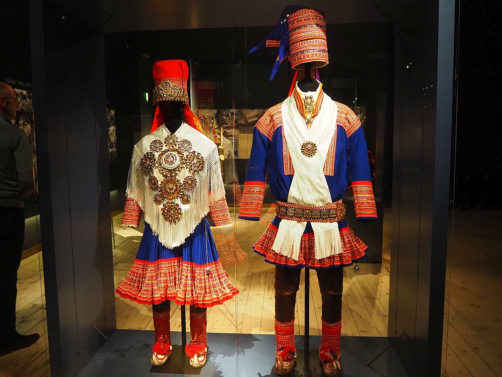 Among the things to do in Rovaniemi is to visit Arktikum. On the right is a woman's outfit, with blue and red embroidered skirt, red tights, a white, very decorated poncho with fringes, and a conical red hat. The mans outfit next to it is in the same colors, but with dark, close-fitting pants and a shorter skirt. Instead of a poncho, a white scarf is around the neck and tucked into a highly decorated belt. The hat is also conical, but more richly embroidered than the woman's hat.