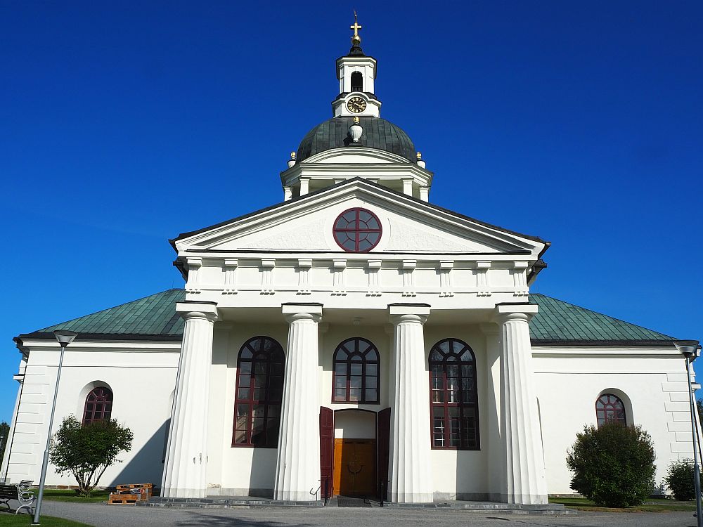 The church has a portico with four very large columns in neoclassical style. The pediment is simple, with a round window in its peak. Above the portico, part of the domed tower is visible: a grey-roofed dome, with a small white steeple on top. A clock is at the base of the steeple and a cross at the top.