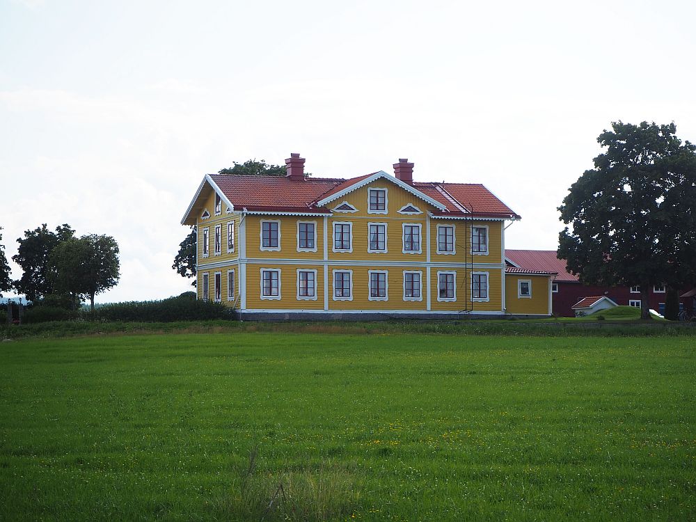 In the foreground, a green field of grass. In the center beyond the filed, a large rectangular house, painted yellow with white trim around the windows. Two stories, with smaller windows under the roof as well. 