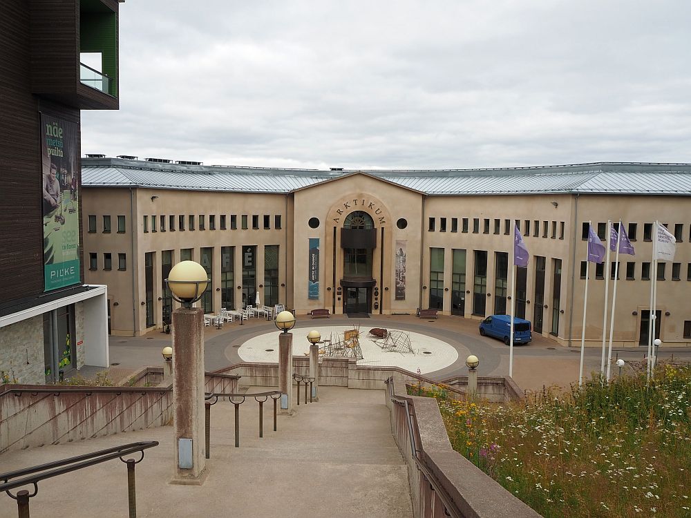 Among the things to do in Rovaniemi is to visit Arktikum. As seen from up a long staircase, the Arkticum building has an arched central entrance with a wing on either side. The building is a light pinkish brown color, and has high, multi-story windows. 