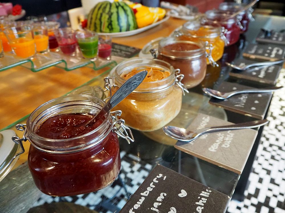 In the foreground, a row of neat containers, each with a spoon and a lable. The nearest says strawberry jam, the second is peanut butter, but there are many more in the row. Behind on a series of small shelves are small glasses of green, red or yellow liquid. 