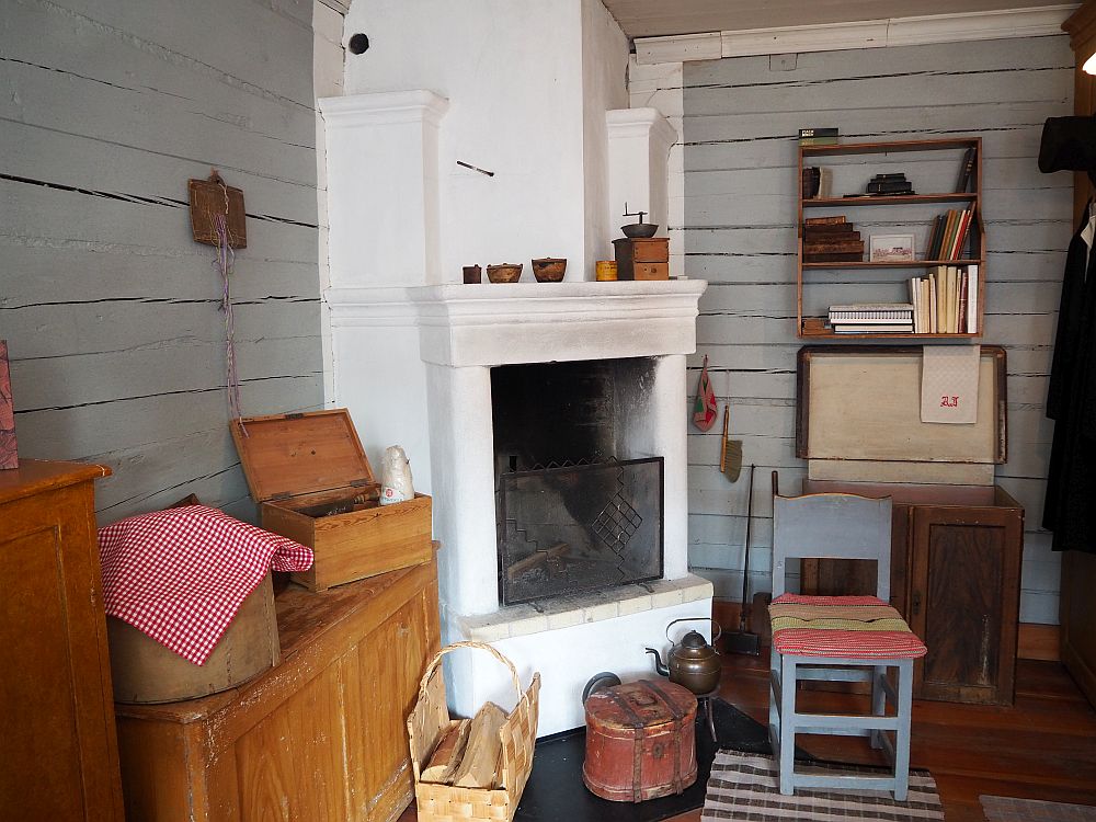 The walls are plain, gray-painted lumber. In the corner is a white painted fireplace. and hearth. On the left is a large chest with a couple of smaller boxes sitting on top of it. In front of the fire is a basket with logs, a round box of some sort, and a tea kettle. Next to the fireplace is a wooden chair with a cushion. Against the wall to the right of the fireplace is another large chest with a small bookshelf mounted on the wall above it.