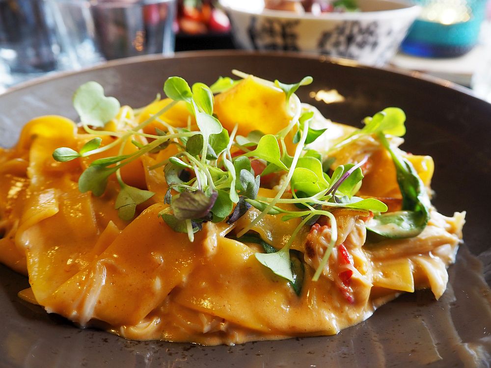 On a dark brown dish, a pile of wide pasta noodles with a creamy sauce and a sprinkling of watercress on top.