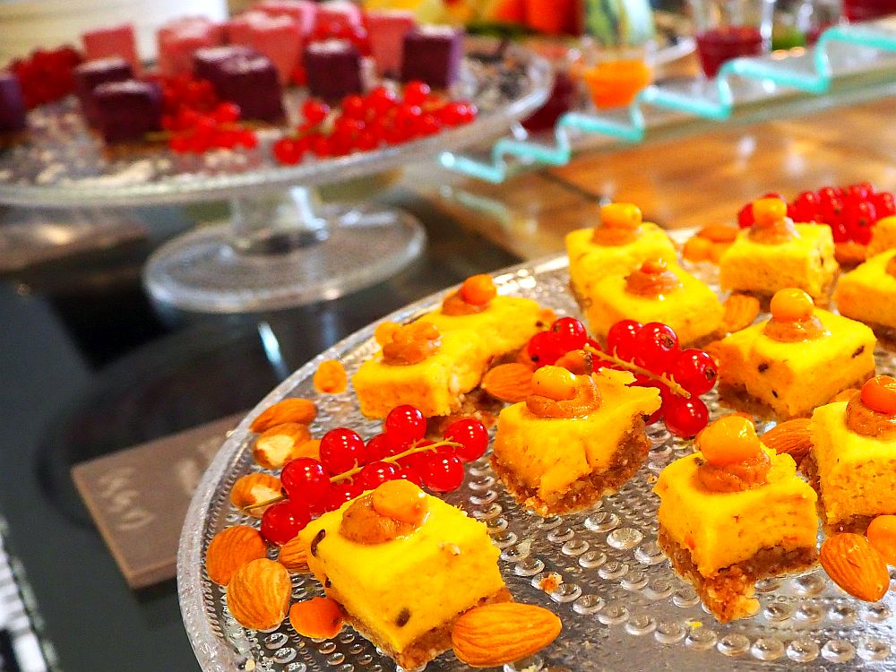 In the foreground, small squares of cake are yellow on what looks like a cookie later. Around them are occasional almonds and redcurrants. In the background, blurry, is another tray with dark brown squares of cake and redcurrants.