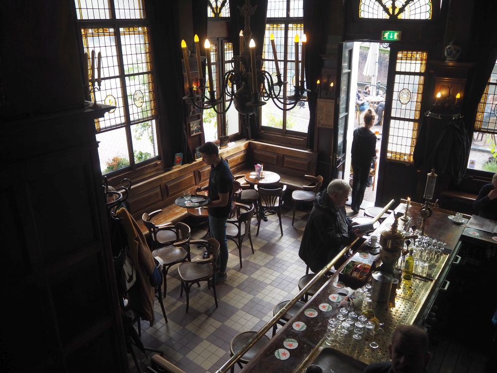 Looking from above down on a small space. Large vertical windows on the left and ahead allow light in. About 3 small round tables are visible with chairs around them and a bench along the left wall. All are brown. the floor is brown tiles. On the right part of the bar is visible with one person sitting on a barstool.