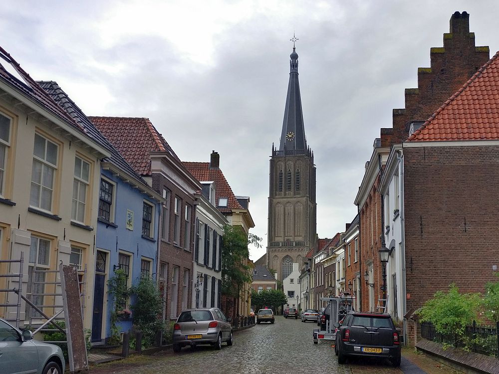 A view down a cobbled street with pretty row houses down each side, some red brick, some painted various colors. At the end of the street, a tall church tower in brown stone.